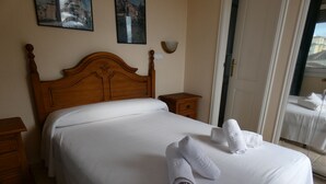 Ein ordentlich bezogenes Bett mit weißen Leinen und aufgerollten Handtüchern. Ein hölzernes Kopfteil, ein Nachttisch mit Lampe und eine Tür zu einem anderen Raum sind zu sehen.