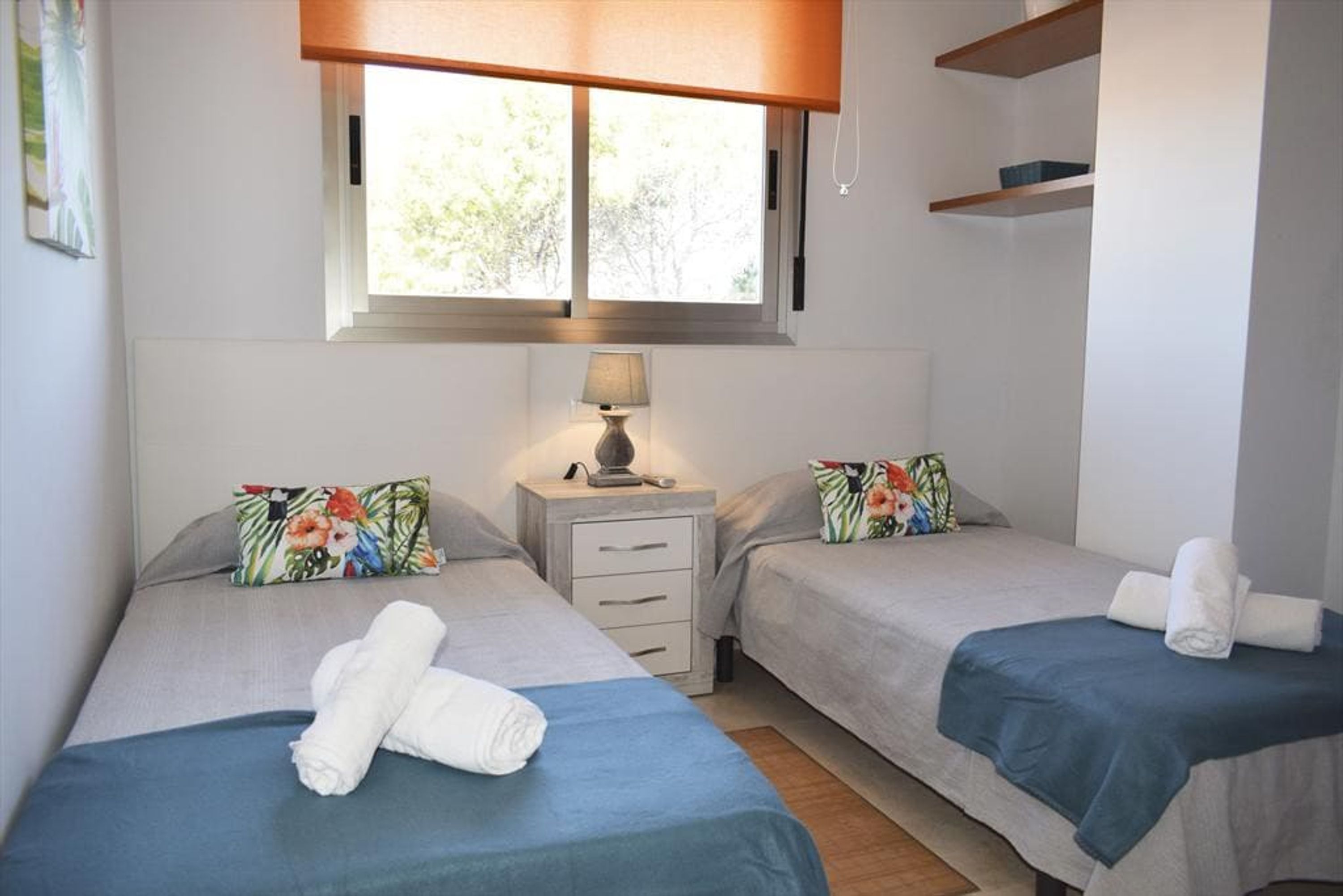 Ein Hotelzimmer mit zwei Betten, jeweils mit blauer Bettdecke und floralen Kissen. Ein Fenster bietet Blick auf Bäume, ein Nachttisch mit Lampe und eine kleine Ablage über den Betten.