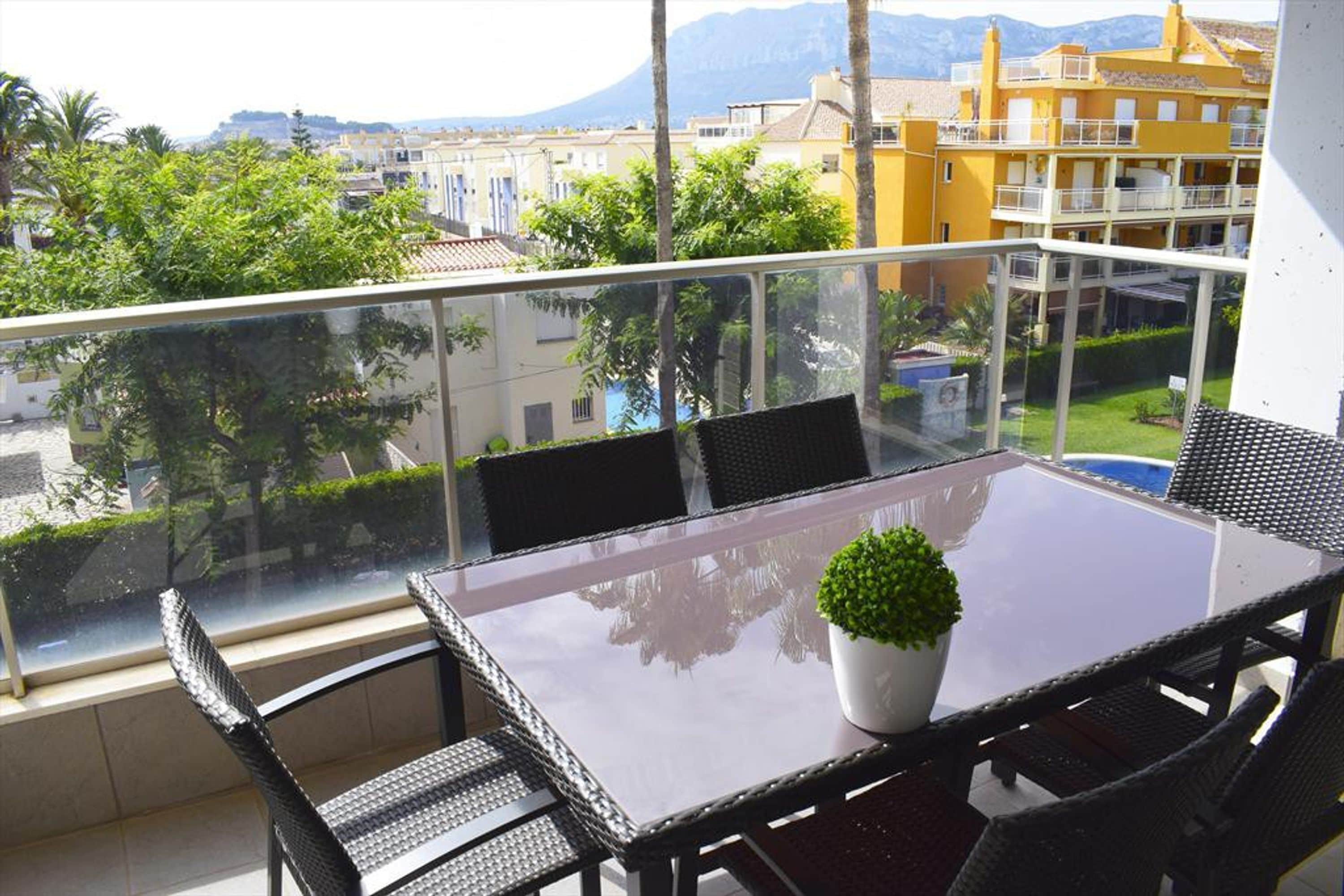 Un balcón con una mesa y sillas de vidrio, una planta en maceta y vistas a edificios y vegetación.