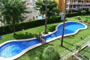 Une piscine avec un pont, entourée de pelouse et d’arbres bien entretenus, et un bâtiment visible au loin.