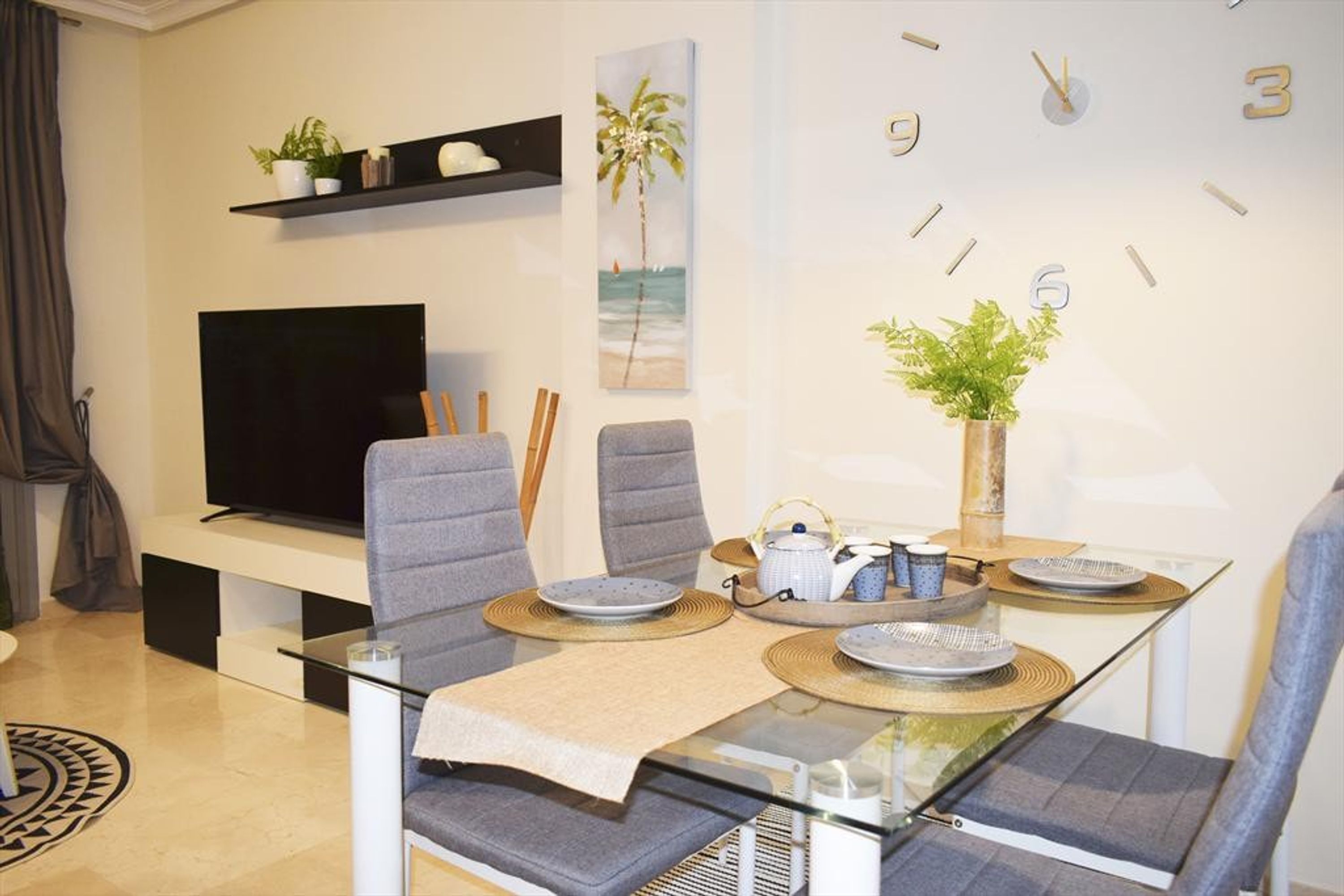 Una sala de estar moderna con un televisor de pantalla plana, sillas tapizadas en gris, una mesa de centro de vidrio y un reloj de pared.