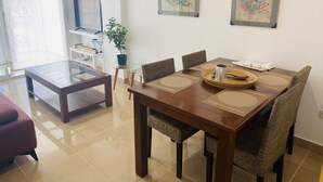 Un comedor con una mesa de madera, sillas de mimbre y una mesa baja de vidrio. Hay dos cuadros en la pared y una planta en maceta sobre una mesita.