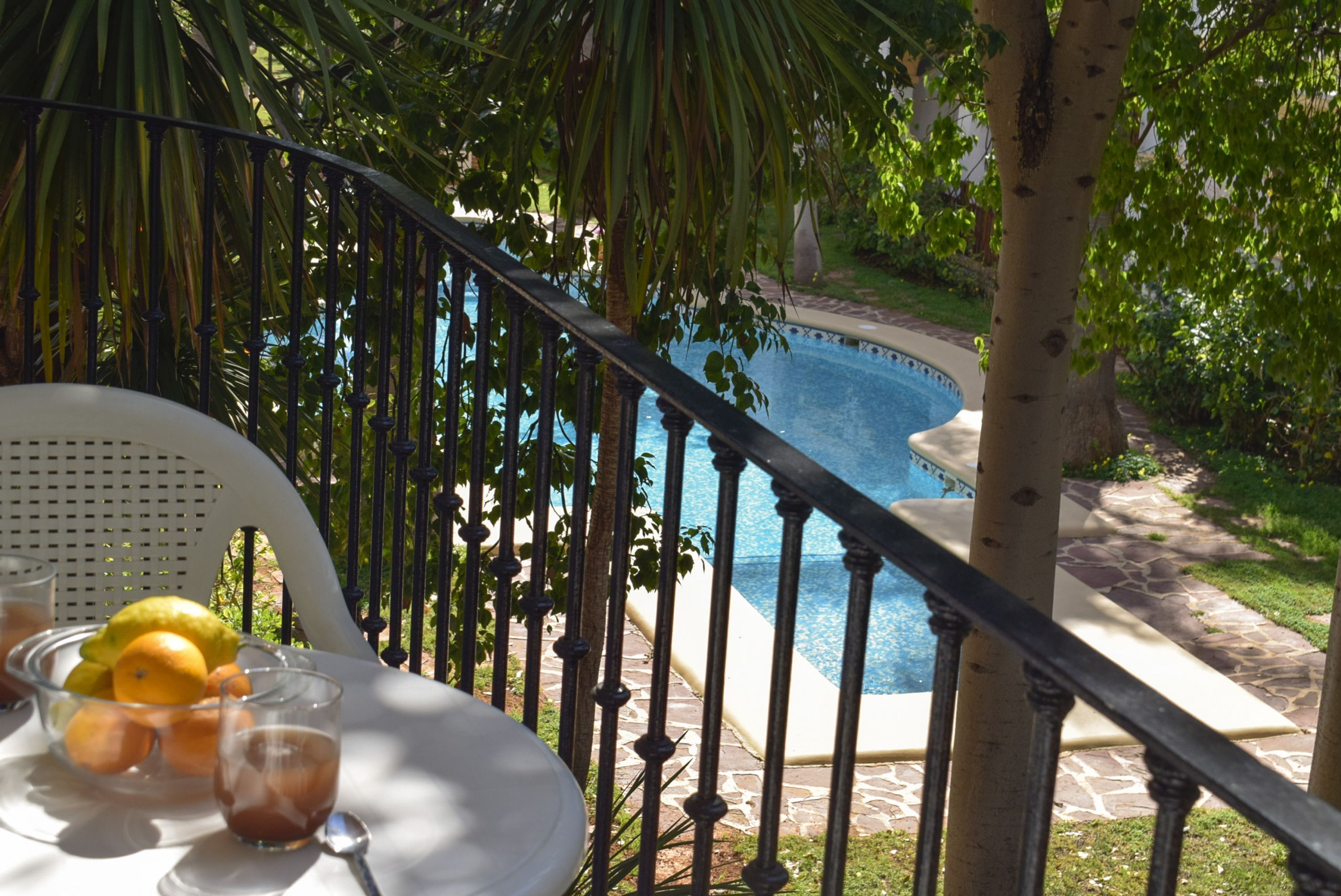Un balcón con vista a la piscina, una mesa con frutas y bebidas, y una silla.