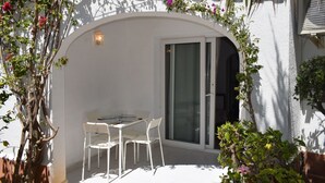 En udendørs terrasse med et bord og stole, en skydedør af glas samt en hvid væg med slyngplanter.