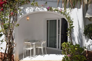 Un patio exterior con mesa y sillas, un ventanal corredero y una pared blanca con plantas trepadoras.