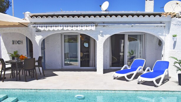 A poolside area with a white building, two blue lounge chairs, and a table with chairs. - Ch3400 in Denia With 3 Bedrooms and 2 Bathrooms (Denia)