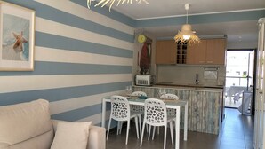 Una stanza con pareti azzurre e bianche a righe, una zona pranzo con sedie bianche, una cucina con mobili in legno e un lampadario al soffitto.