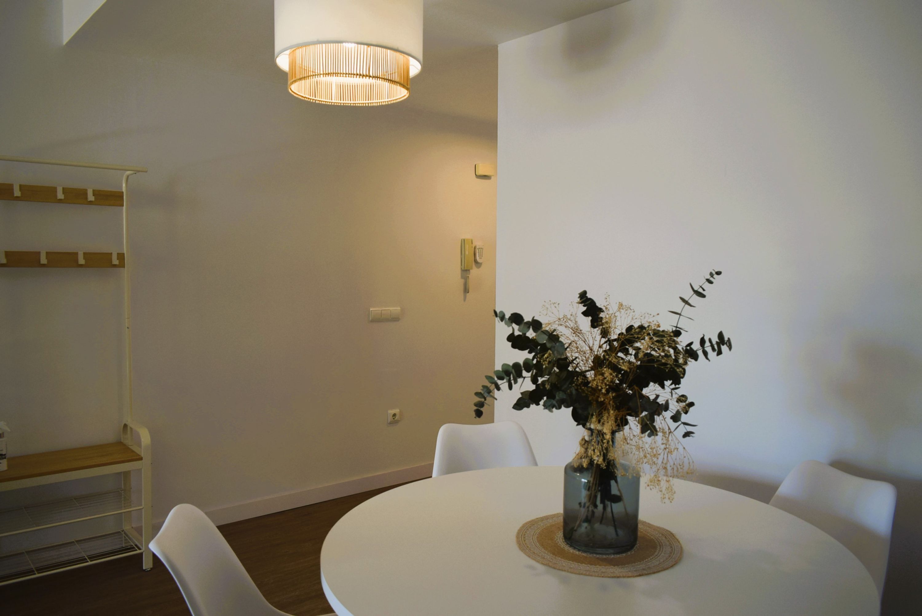 A modern dining area with a round white table, white chairs, a vase with dried flowers, and a hanging light fixture.
