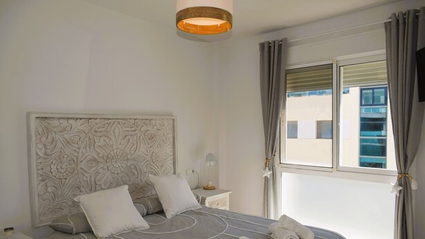 Ein ordentlich bezogenes Bett mit einem gemusterten Kopfteil, ein Nachttisch mit einer Lampe und ein Fenster mit Vorhängen.