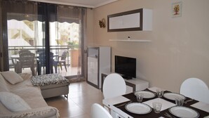 Ein modernes Wohnzimmer mit Sofa, einem Esstisch für vier Personen, einem Fernseher und einem Balkon mit Vorhängen.