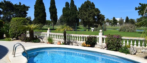 A swimming pool with a stone edge, surrounded by a white balustrade, with a view of a golf course and tall trees in the background.