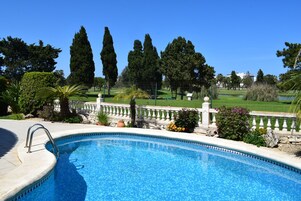 A swimming pool with a stone edge, surrounded by a white balustrade, with a view of a golf course and tall trees in the background.