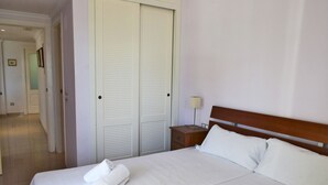 Egy gondosan megterített ágy fehér ágyneművel és egy feltekert törölközővel, egy fa éjjeliszekrény lámpával, és egy zárt, fehér szekrény.