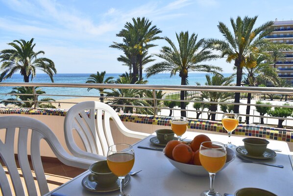 A table with a bowl of fruit and glasses of orange juice, overlooking a beach with palm trees and a clear sky. - Ap3208 in Denia With 3 Bedrooms and 2 Bathrooms (Denia)