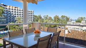 Un balcon avec une table et des chaises en verre, un saladier rempli d’oranges et une vue sur des palmiers et des immeubles.