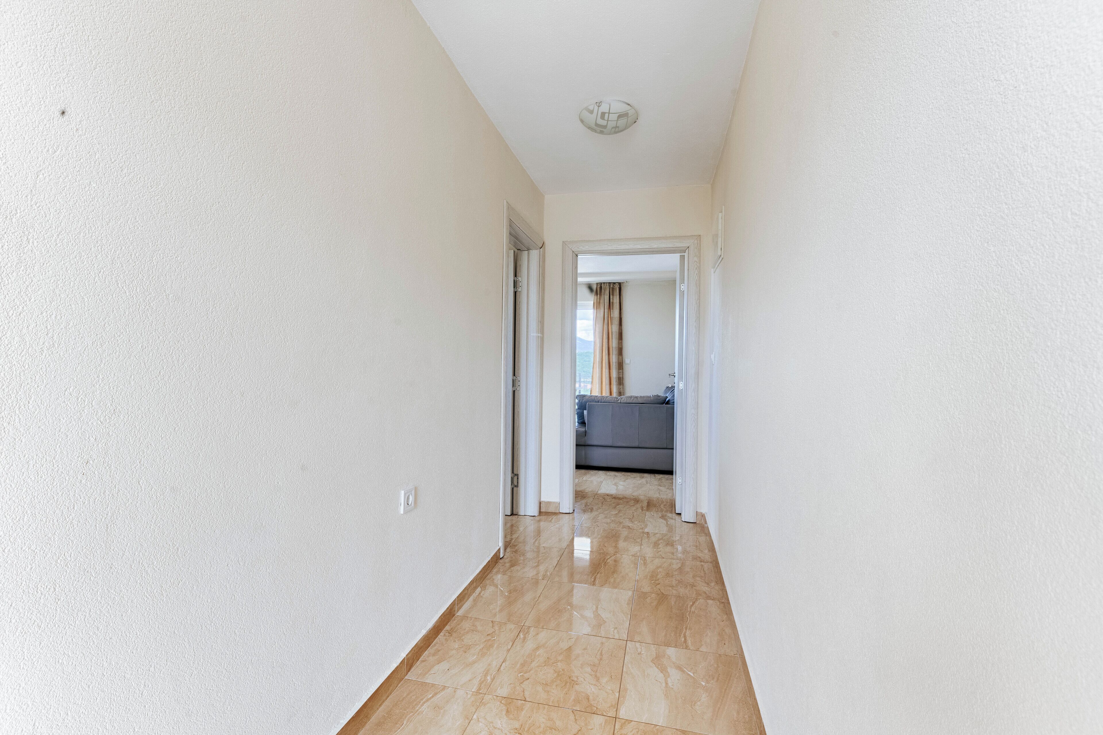 Un pasillo con paredes blancas y suelo de baldosas que conduce a una habitación con un sofá gris.