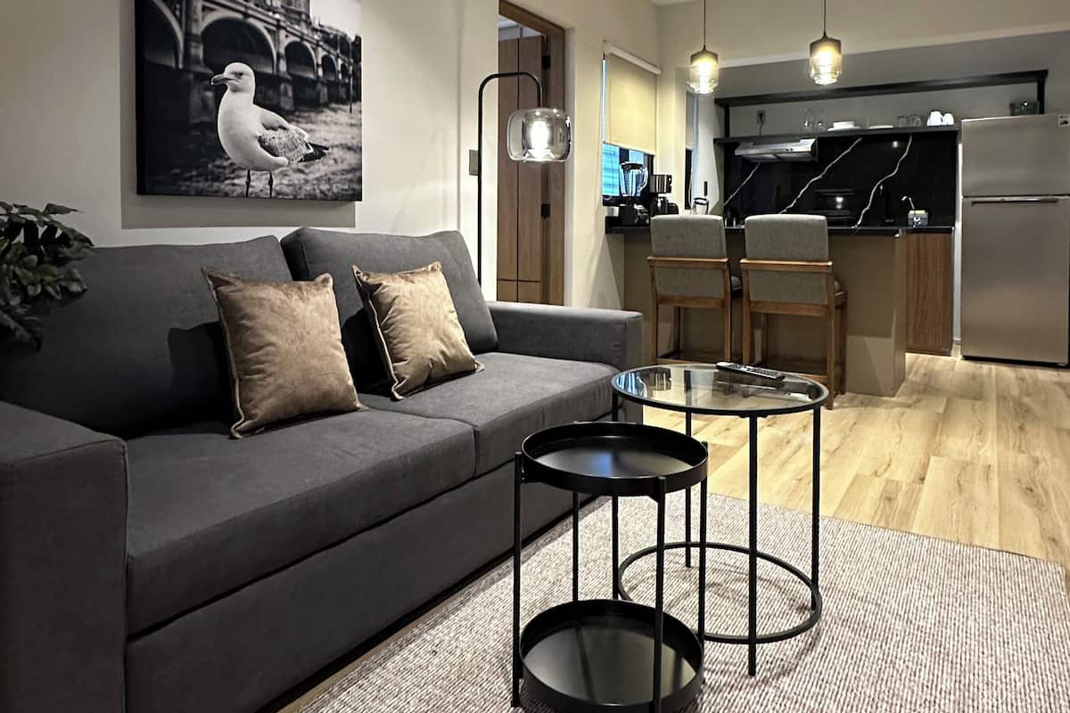 A modern living room with a grey sofa, two black side tables, and a painting of a bridge.