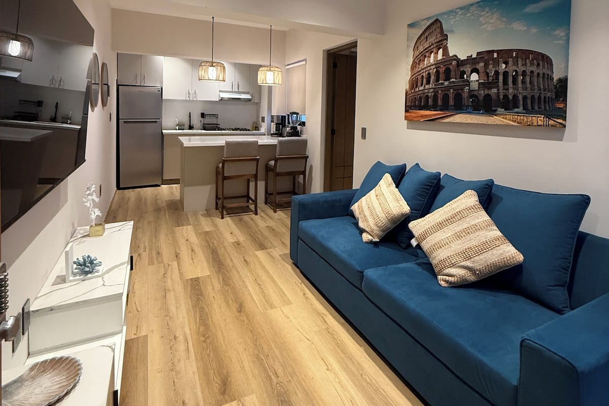 A modern living room with a blue sofa, wooden flooring, and a kitchen in the background.