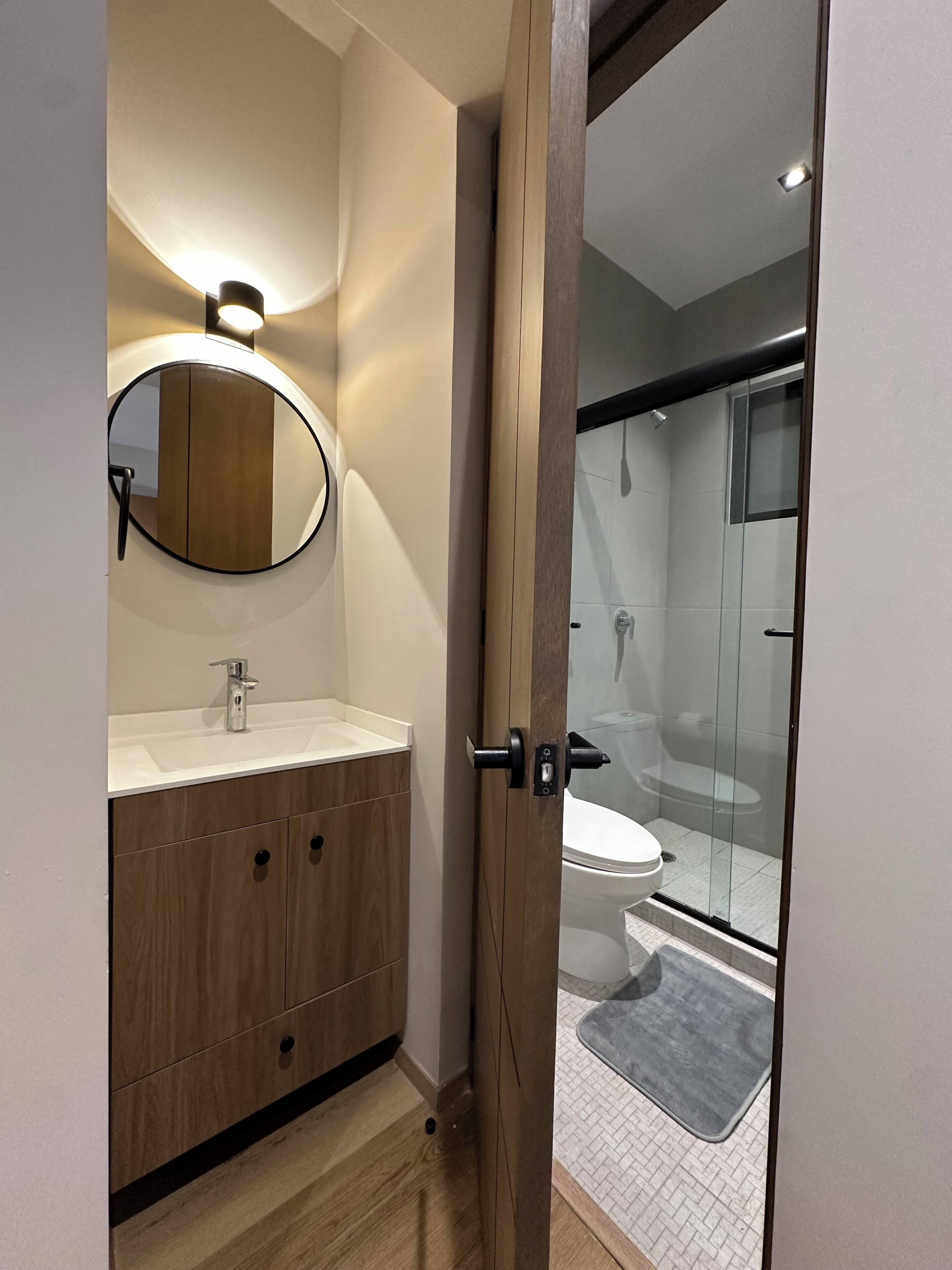 A modern bathroom with a wooden vanity, a round mirror, and a glass shower enclosure.