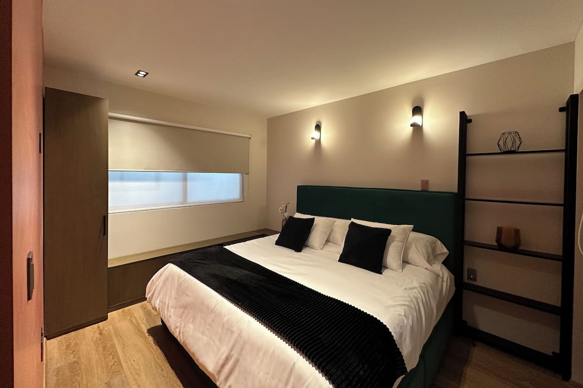 A bedroom with a bed, a window with blinds, a headboard, wall-mounted lights, and a shelf with decorative items.