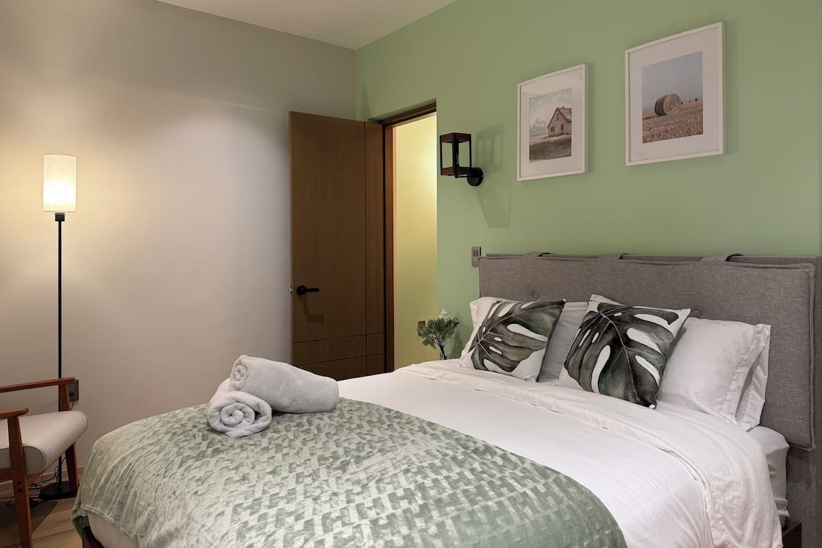 A neatly made bed with a patterned bedspread, a rolled towel, and a bedside lamp. There are two framed pictures on the wall above the bed.