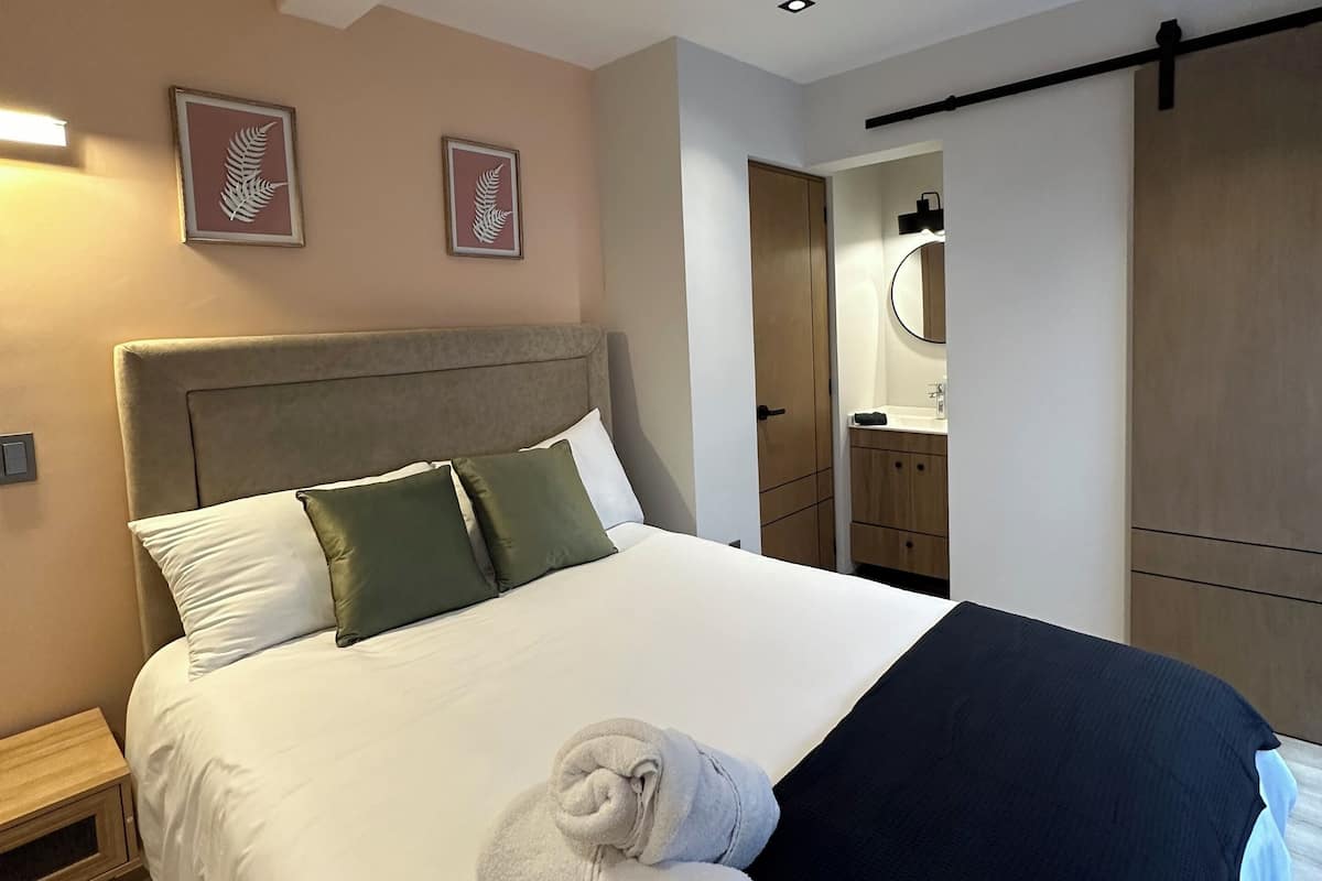 A neatly made bed with white and green pillows, a black blanket, and a rolled towel. There are framed leaf designs on the wall, a wooden wardrobe, and a small bedside table.
