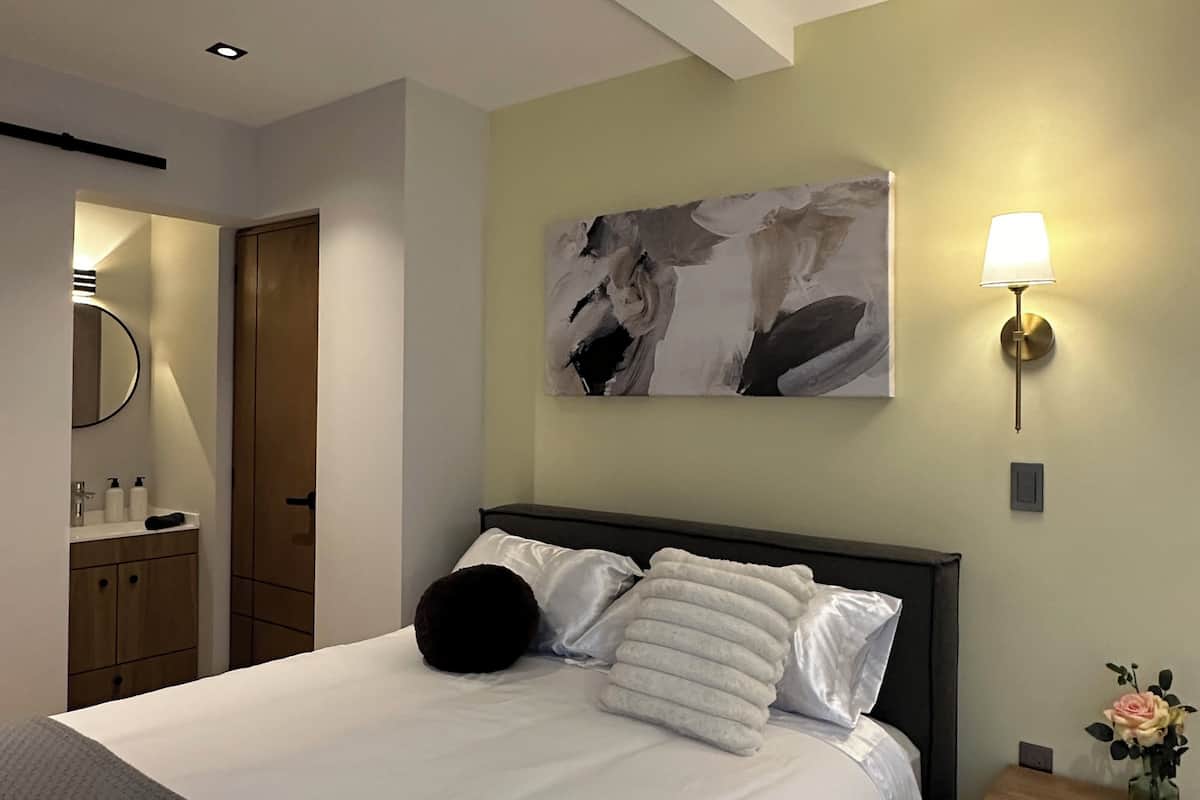 A modern hotel room with a large bed, a bedside table with a lamp, a wall-mounted artwork, and a bathroom visible through an open door.