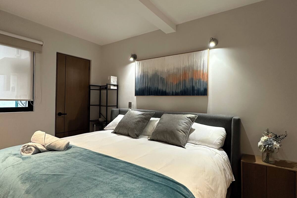 A modern bedroom with a large bed, a headboard with integrated lighting, a wall-mounted abstract painting, and a bedside table with a vase.