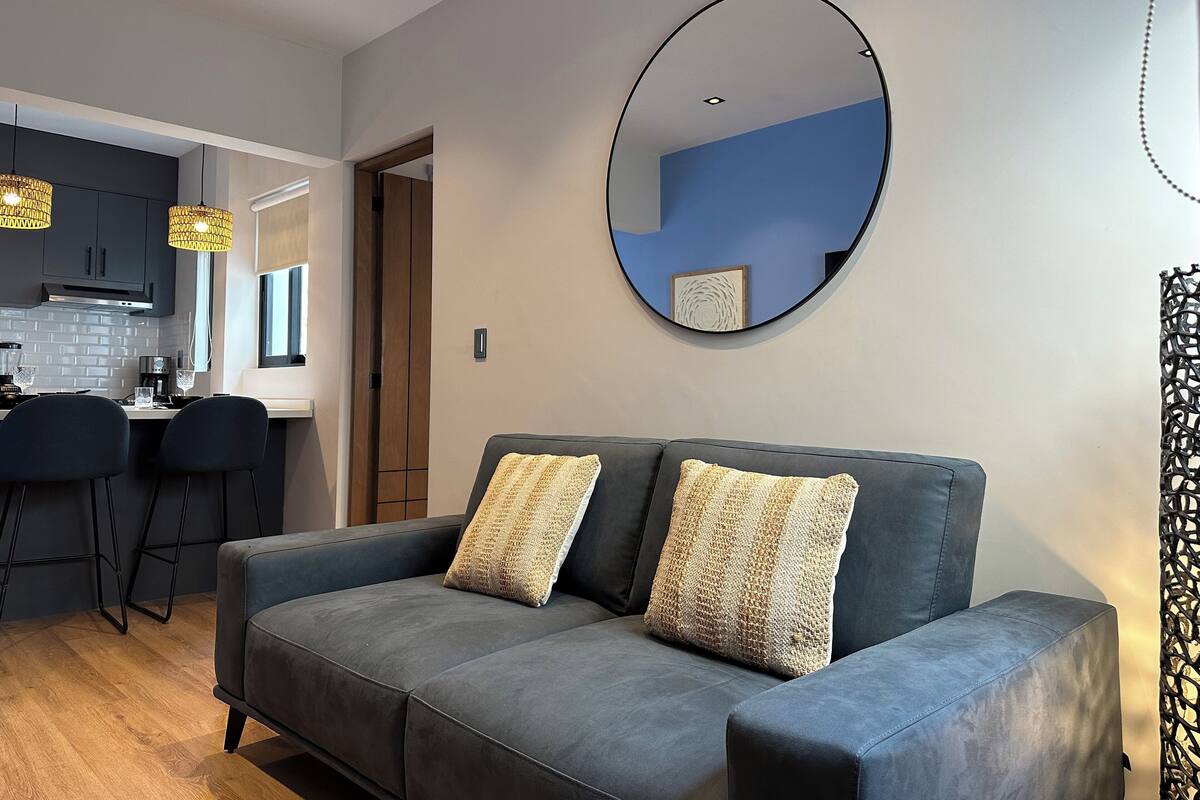 A modern living room with a grey sofa, two patterned cushions, a round mirror on the wall, and a kitchen area in the background.