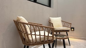 Deux fauteuils en osier avec des coussins, placés contre un mur percé d’une grande fenêtre.