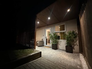 A nighttime view of a house's exterior with a paved patio, a door leading inside, and two potted plants. - Cosy, self-contained guest house (London)