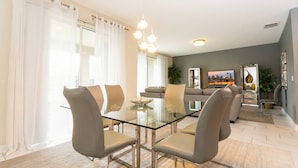Ett modernt matområde med ett glasbord och stolar, en soffa i bakgrunden och ett stort fönster med gardiner.