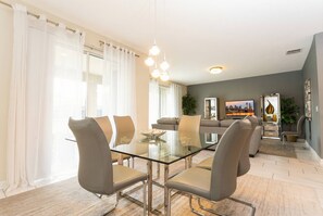 A modern dining area with a glass table and chairs, a sofa in the background, and a large window with curtains. - Luxury 6BR Resort Villa Near Disney (Kissimmee)