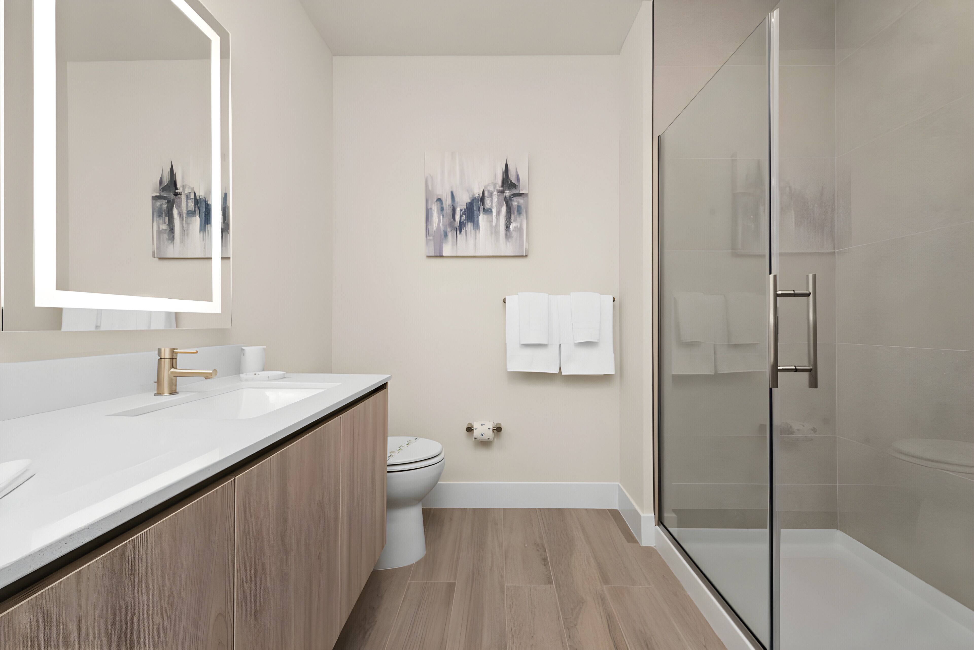 A modern bathroom with a wooden vanity, a glass shower enclosure, and two abstract paintings on the walls.
