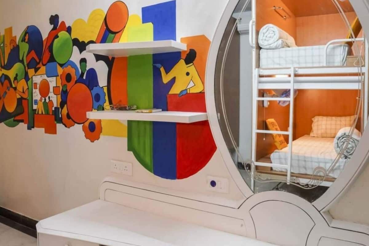 Un lit superposé avec une fenêtre ronde, une commode blanche à tiroirs et une œuvre d’art murale abstraite et colorée.