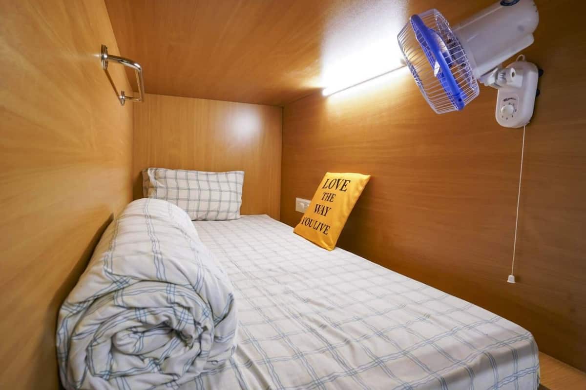 Une petite pièce agréable et confortable, avec un lit, un ventilateur fixé au mur et un coussin décoré d’une citation inspirante.