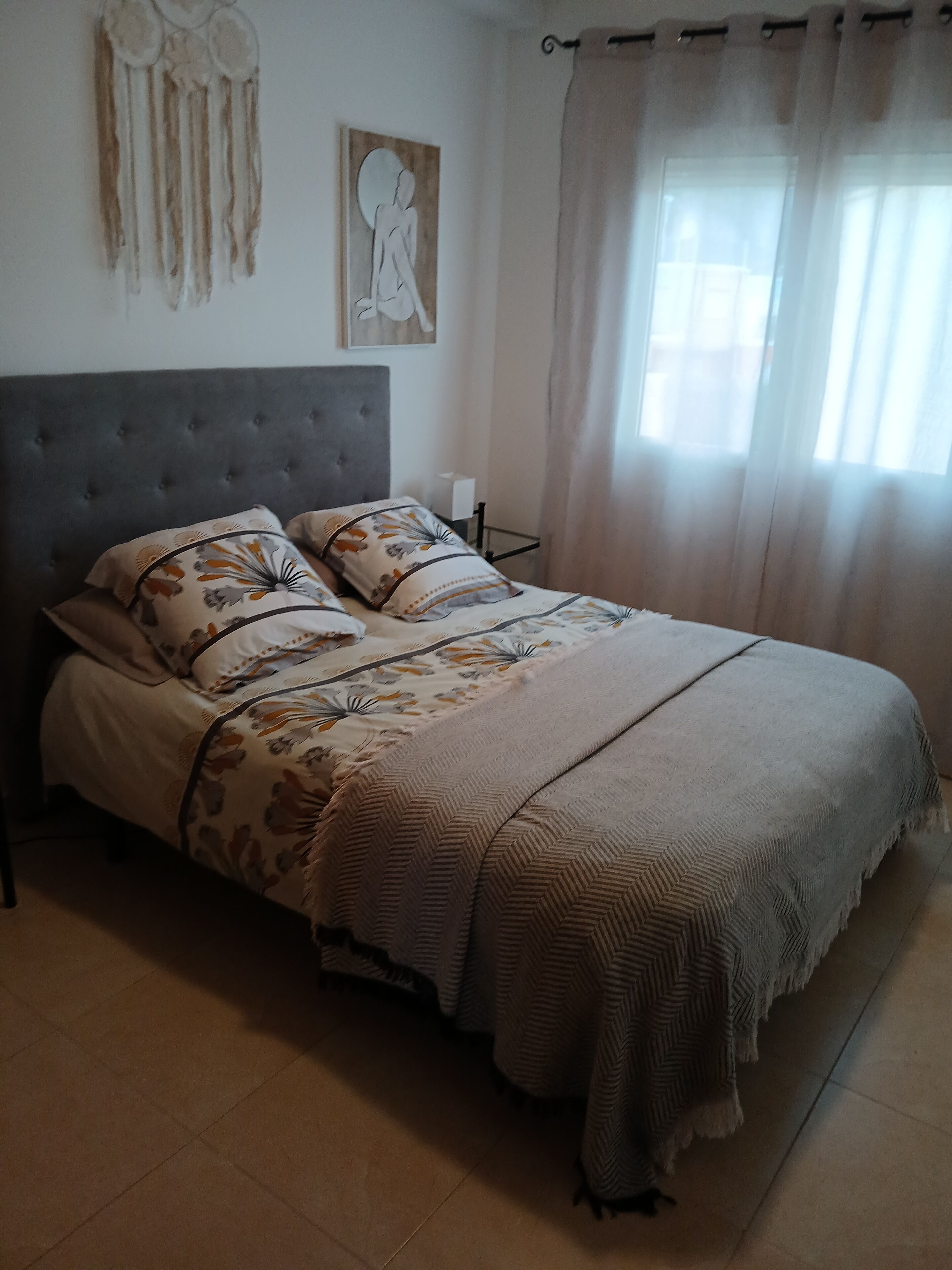 A bedroom with a tufted headboard, floral patterned bedding, and a large window with sheer curtains.