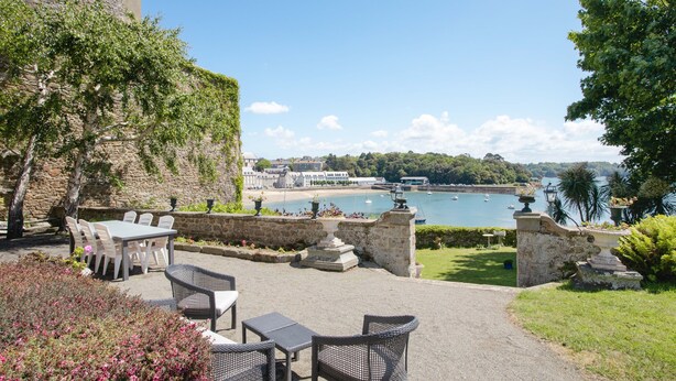 Eine Terrasse im Freien mit Steinmauern, einem Tisch und Stühlen, mit Blick auf eine Bucht mit Booten und einer fernen Küste.