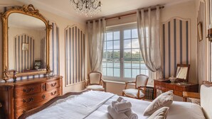 Ein Hotelzimmer mit einem großen Bett, einer Holzkombination, einem Spiegel, zwei Stühlen, einem Fenster mit Vorhängen und einer Chandelier.