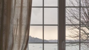 Ein Zimmer mit großem Fenster mit Blick auf ein Gewässer und Berge. Ein Sessel und eine Heizung sind sichtbar.