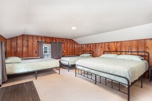 A room with three metal beds, wooden paneling on the walls, a window with curtains, and a carpeted floor.