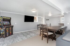 A room with a dining table, chairs, a television, and a video game console.