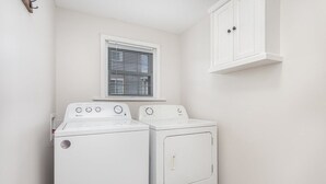 Une buanderie avec deux machines à laver blanches, une fenêtre avec des stores et un meuble blanc.