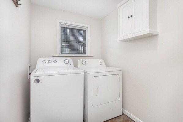 A laundry room with two white washing machines, a window with blinds, and a white cabinet.