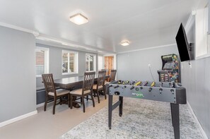 A room with a foosball table, a dining table with chairs, and a television mounted on the wall.