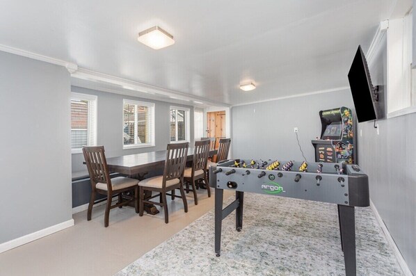 A room with a foosball table, a dining table with chairs, and a television mounted on the wall.