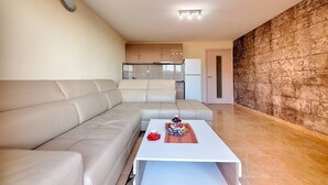 A modern living room with a beige sofa, a white coffee table, and a kitchen in the background. - Apartman ZIS 5D (Opština Bar)