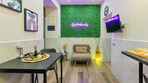 Une pièce moderne avec un mur vert, un néon, un téléviseur à écran plat, une table à manger avec de la nourriture, un fauteuil confortable et une petite table d’appoint.