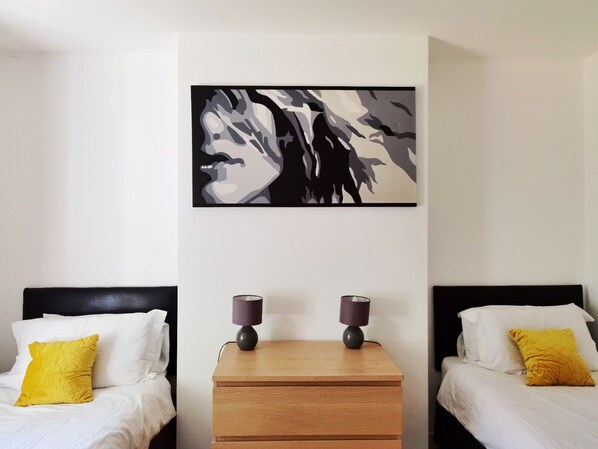 A hotel room with two beds, each with white bedding and a yellow pillow. A wooden nightstand with two lamps is centered between the beds. A large abstract painting is hung on the wall above the nightstand. - Perry Road (Nottingham)
