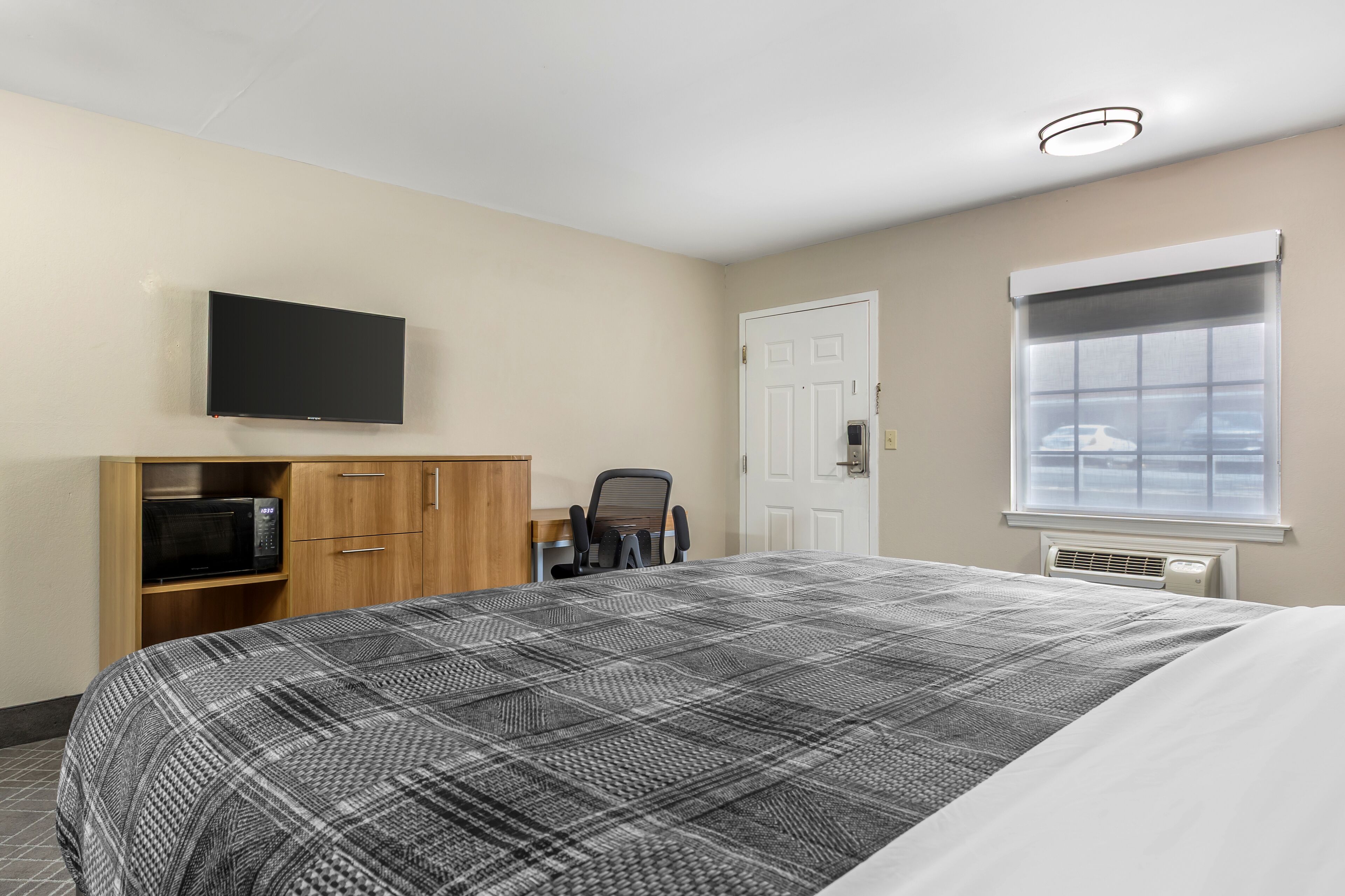 Habitación de hotel con cama, televisor montado en la pared, armario de madera, silla y ventana con vistas.